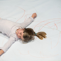 Zdjęcie dziewczynki leżącej z rozłożonymi rękami na dużym arkuszu papieru popisanego kredkami.