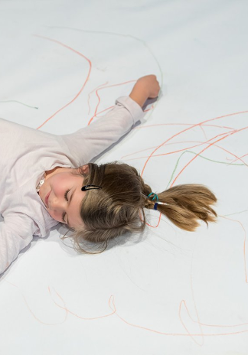 Zdjęcie dziewczynki leżącej z rozłożonymi rękami na dużym arkuszu papieru popisanego kredkami.