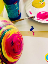 Kolorowe zdjęcie żółtego balona częściowo pomalowanego w kolorowe okręgi. Na drugim planie papierowy talerz z trzema kolorowymi plamami, o talerz oparty jest pędzelek.