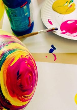 Kolorowe zdjęcie żółtego balona częściowo pomalowanego w kolorowe okręgi. Na drugim planie papierowy talerz z trzema kolorowymi plamami, o talerz oparty jest pędzelek.