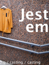 Fragment ściany z poręczą, na ścianie wisi pomarańczowa bluzka z metką od ceny. Na środku napis w dwóch linijkach "jest em".