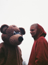 Zdjęcie młodego chłopaka w bordowej bluzie z kapturem, który stoi obok pluszowego misia równego mu wzrostem.