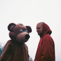 Zdjęcie młodego chłopaka w bordowej bluzie z kapturem, który stoi obok pluszowego misia równego mu wzrostem.