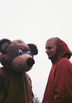 Zdjęcie młodego chłopaka w bordowej bluzie z kapturem, który stoi obok pluszowego misia równego mu wzrostem.