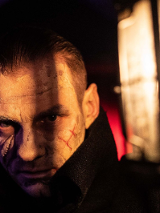 Twarz mężczyzny w półmroku, na którą częściowo pada światło. Na twarzy widoczne blizny.