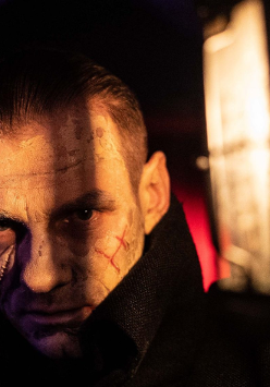 Twarz mężczyzny w półmroku, na którą częściowo pada światło. Na twarzy widoczne blizny.