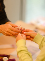 Dziecko, które wspólnie z dorosłą osobą trzyma w dloniach kawałki czerwonego sznurka. Twarz dziecka widoczna jest tylko częściowo, twarz osoby dorosłej nie jest widoczna.