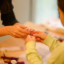 Dziecko, które wspólnie z dorosłą osobą trzyma w dloniach kawałki czerwonego sznurka. Twarz dziecka widoczna jest tylko częściowo, twarz osoby dorosłej nie jest widoczna.