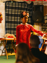 Trzy kobiety na scenie, są podobnie ubrane, każda z nich ma kokardę we włosach. W tle przedmioty codziennego użytku, w tym stacjonarny telefon na stoliku.