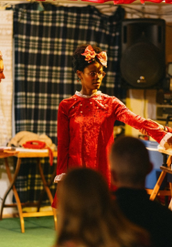 Trzy kobiety na scenie, są podobnie ubrane, każda z nich ma kokardę we włosach. W tle przedmioty codziennego użytku, w tym stacjonarny telefon na stoliku.