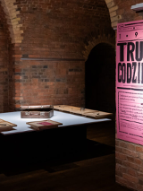 Wnętrze sali wystawowej w ciemnym piwnicznym pomieszczeniu. Na ścianie oświetlony plakat wystawy, w tle stół, na którym leży kilka przedmiotów, m.in. mały kuferek.