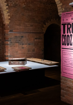 Wnętrze sali wystawowej w ciemnym piwnicznym pomieszczeniu. Na ścianie oświetlony plakat wystawy, w tle stół, na którym leży kilka przedmiotów, m.in. mały kuferek.