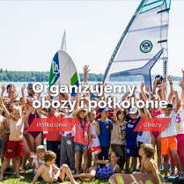 Dzieci i trenerzy w strojach kapielowych i deskami windserfingowymi pozujący do zdjęcia grupowego na trawie. W tle jezioro i żagle.