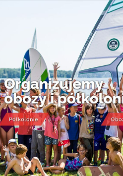 Dzieci i trenerzy w strojach kapielowych i deskami windserfingowymi pozujący do zdjęcia grupowego na trawie. W tle jezioro i żagle.