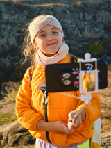 Dziewczynka w wełnianej opasce i pomarańczowej kurtce robi sobie zdjęcie na łące przy użyciu selfie stick'a.