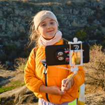 Dziewczynka w wełnianej opasce i pomarańczowej kurtce robi sobie zdjęcie na łące przy użyciu selfie stick'a.