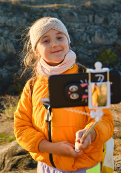 Dziewczynka w wełnianej opasce i pomarańczowej kurtce robi sobie zdjęcie na łące przy użyciu selfie stick'a.