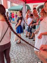 Kobieta zaplata warkocz dziewczynie przed nią. Dookła inni ludzie oraz stragany.