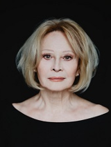 Zdjęcie kobiety w jasnych włosach, ubranej na czarno, na czarnym tle. Kobieta patrzy prosto w obiektyw.