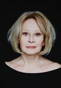 Zdjęcie kobiety w jasnych włosach, ubranej na czarno, na czarnym tle. Kobieta patrzy prosto w obiektyw.