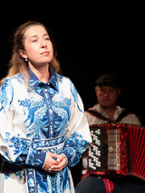 Zdjęcie kobiety w biało-niebieskiej sukience, która patrzy w dal. W tle mężczyzna grający na akordeonie.