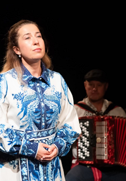 Zdjęcie kobiety w biało-niebieskiej sukience, która patrzy w dal. W tle mężczyzna grający na akordeonie.