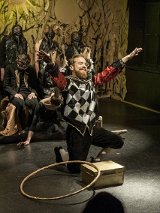 na pierwszym planie mężczyna klęczący na jedno kolano, przed nim stoi pudełko i leży hulahop. Mężczyzna ma kolorowy strój: czarne spodnie do kolan, białe kolanówki, czerwoną koszulę, kamizelkę w biało-czarną szachownicę. W tle kilka postaci ubranych na czarno w różnych pozach.