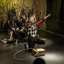 na pierwszym planie mężczyna klęczący na jedno kolano, przed nim stoi pudełko i leży hulahop. Mężczyzna ma kolorowy strój: czarne spodnie do kolan, białe kolanówki, czerwoną koszulę, kamizelkę w biało-czarną szachownicę. W tle kilka postaci ubranych na czarno w różnych pozach.