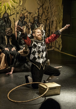 na pierwszym planie mężczyna klęczący na jedno kolano, przed nim stoi pudełko i leży hulahop. Mężczyzna ma kolorowy strój: czarne spodnie do kolan, białe kolanówki, czerwoną koszulę, kamizelkę w biało-czarną szachownicę. W tle kilka postaci ubranych na czarno w różnych pozach.