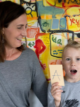 Kobieta w szarym swetrze odwórcona w stronę dziecka siedzącego po prawej uśmiecha się do niego. Zadowolony chłopiec trzyma kartkę z literą "A".