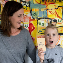 Kobieta w szarym swetrze odwórcona w stronę dziecka siedzącego po prawej uśmiecha się do niego. Zadowolony chłopiec trzyma kartkę z literą "A".