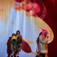 Dwie kobiety na scenie ubrane w futra i trzymające mikrofony. Oświetlone czerwono-brzoskwiniowymi światłami.