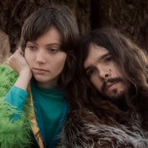 Kobieta i mężczyzna opierający się o siebie ze wzrokiem w daleką przestrzeń. W tle pień drzewa. Para ubrana jest w sztuczne futra.