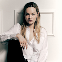 Zdjęcie Michaliny łabacz - młodej kobiety, w białej bluzce i czarnych spodniach, siedzacej pod ścianą, z jedną nogą zgiętą w kolanie.