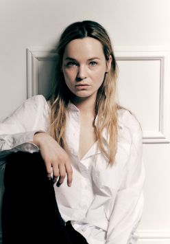 Zdjęcie Michaliny łabacz - młodej kobiety, w białej bluzce i czarnych spodniach, siedzacej pod ścianą, z jedną nogą zgiętą w kolanie.