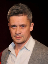 Zdjęcie uśmiechniętego mężczyzny w średnim wieku, ubranego w jasną koszulę i szary sweter