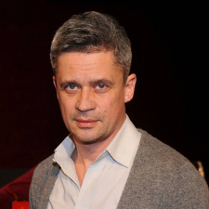 Zdjęcie uśmiechniętego mężczyzny w średnim wieku, ubranego w jasną koszulę i szary sweter