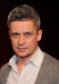 Zdjęcie uśmiechniętego mężczyzny w średnim wieku, ubranego w jasną koszulę i szary sweter