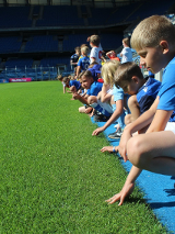 Zdjęcie przedstawiuczestników półkolonii, którzy w strojach piłkarskich siedzą przy boisku i dotykają trawy.