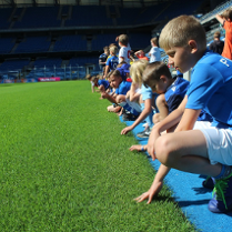 Zdjęcie przedstawiuczestników półkolonii, którzy w strojach piłkarskich siedzą przy boisku i dotykają trawy.