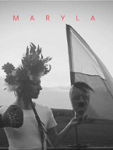 Czarno-białe zdjęcie kobiety z kwiecistym wiankiem na głowie, która w jednej ręce trzyma rzeźbę - głowę mężczyzny, a na drugiej ręce ma rękawicę bokserską w pozie do uderzenia. Za kobietą flaga, w tle otwarta przestrzeń.