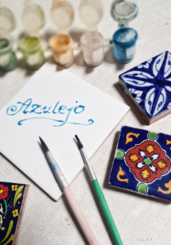 Na zdjęciu znajdują się 4 kafelki w różnych rozmiarach, na jednym widnieje napis "Azulejo", a pozostałe trzy są pomalowane. Na zdjęciu znajdują się również farbki oraz dwa pędzle.