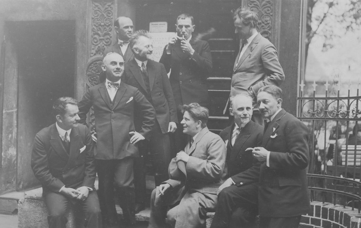 Adam Ballenstedt pierwszy z prawej (siedzi na murku) Zdjęcie sprzed 100 lat, grupa mężczyzn siedzi, stoi, rozmawiają palą