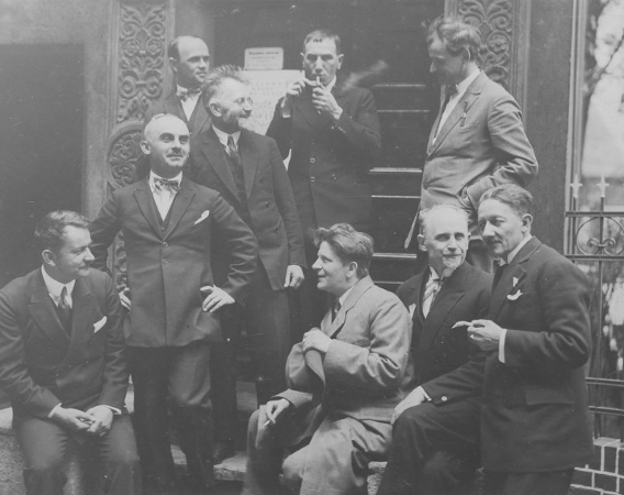 Adam Ballenstedt pierwszy z prawej (siedzi na murku) Zdjęcie sprzed 100 lat, grupa mężczyzn siedzi, stoi, rozmawiają palą