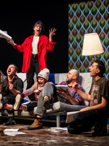 na kanapie na scenie siedzi kilka osób, niektórzy trzymają w rękach otwarte książki. Za nimi stoi kobieta z otwartą książką w ręce, mocno gestykuluje