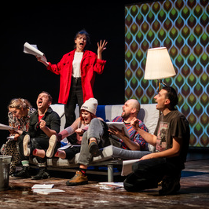 na kanapie na scenie siedzi kilka osób, niektórzy trzymają w rękach otwarte książki. Za nimi stoi kobieta z otwartą książką w ręce, mocno gestykuluje