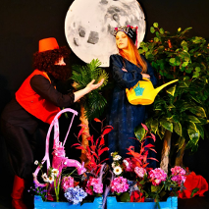 Zdjęcie przedstawie fragment ze spektaklu. Na zdjęciu znajdują się dwie osoby, między dekoracyjnymi roślinami. W tle widac księżyć na czarnym materiale.