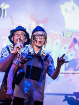 Na zdjęciu znajdują się dwie osoby, które na fioletowym tle ścian i świateł robią nietypowe pozy oraz miny.