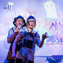 Na zdjęciu znajdują się dwie osoby, które na fioletowym tle ścian i świateł robią nietypowe pozy oraz miny.