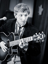 zdjecie wykonawcy - mężczyzna stojący na scenie przy mikrofonie, grający na gitarze